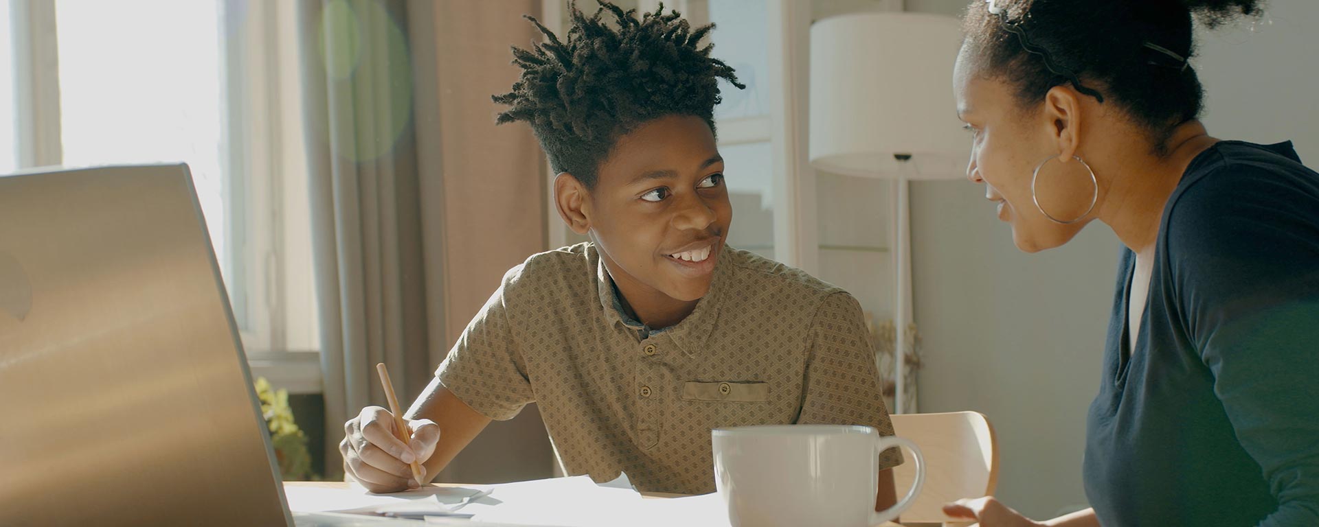 Image of a student and mother. The student is an adolescent black youth with curly black hair. They are writing with a pencil. His mother is also at the table in a green shirt and is looking at them.