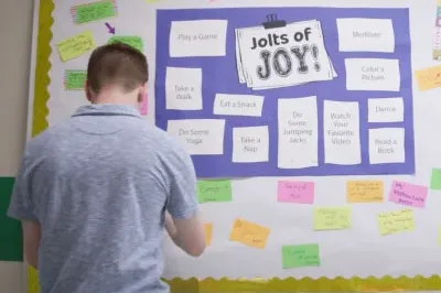 2.15.23_JoJBulletinBoard White student in a blue shirt putting a note onto a bulletin board in a school hallway that has a "Jolts of Joy" poster on it.