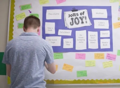 2.15.23_JoJBulletinBoard White student in a blue shirt putting a note onto a bulletin board in a school hallway that has a "Jolts of Joy" poster on it.