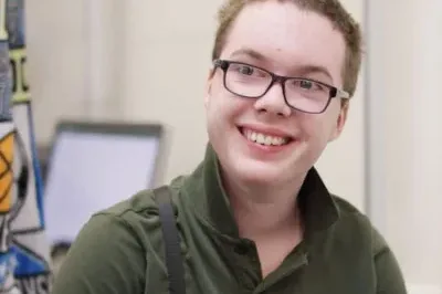 3.3.23_SpreadingPosP Autistic white teenage boy with a green polo and glasses smiling in his classroom.