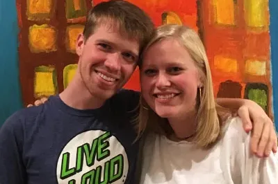 Avery, and white woman with blonde hair wearing a white shirt, and Tyler, a white autistic man with brown hair wearing a blue shirt, smiling and standing in front of one of Tyler's paintings of yellow and orange buildings.
