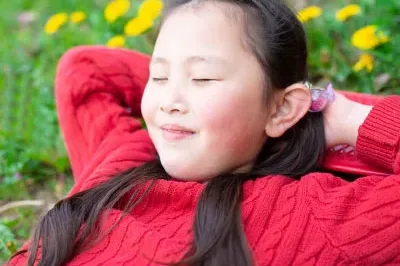 A content young Asian child with brown hair wearing a red sweater lays in a field with her eyes closed.