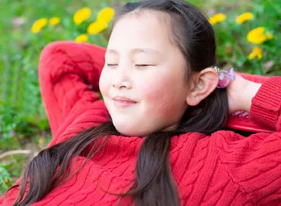 4.28.23_GratitudeGirl_ A content young Asian child with brown hair wearing a red sweater lays in a field with her eyes closed.