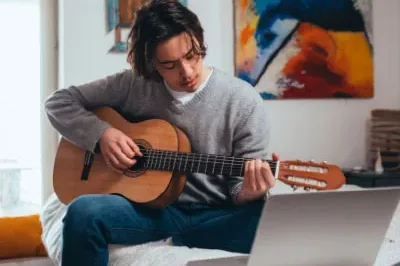 5.11.23_PERMA+Guitar A teenaged boy wearing a grey sweater learning to play guitar