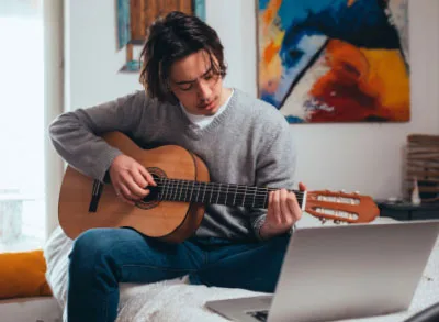 5.11.23_PERMA+Guitar A teenaged boy wearing a grey sweater learning to play guitar