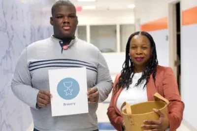 Strength Spotting Barnel and Gloria stand in a school hallway while Strength Spotting around their school. Barnel is holding a sign with the Character Strengths icon for Bravery.