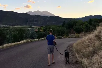 7.13.23_NatureBenefits A man walks down a country road with mountains in the distance. He is facing away from the camera and is walking a dog at sunset.