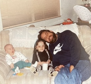 A young Nicole sits between her baby brother and her father on a couch.