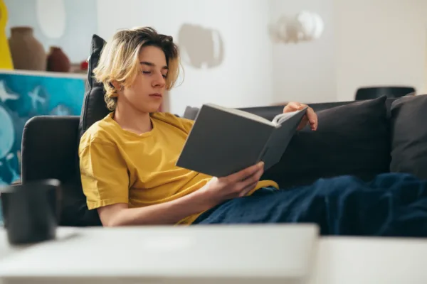 teenager boy reading book at home Teenage boy with blonde hair and a yellow t-shirt sitting on a couch reading a book