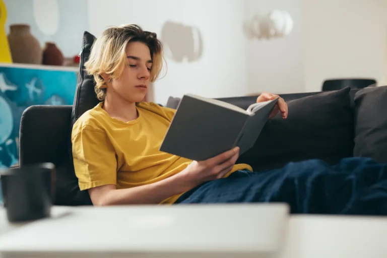 teenager boy reading book at home Teenage boy with blonde hair and a yellow t-shirt sitting on a couch reading a book