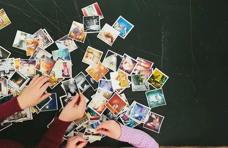 Several colorful photos are spread out over a black surface. Two pairs of small hands are picking up the photos.