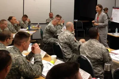 11.10.23_KC_VeteransDay White woman speaking wearing a gray pant suite speaking in front of soldiers sitting in chairs at desks.