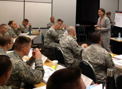 11.10.23_KC_VeteransDay White woman speaking wearing a gray pant suite speaking in front of soldiers sitting in chairs at desks.