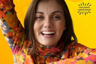 400×293 WW_TAL Tal Anderson is smiling and wearing a colorful flowery turtle neck and holding up her arms against a yellow background.