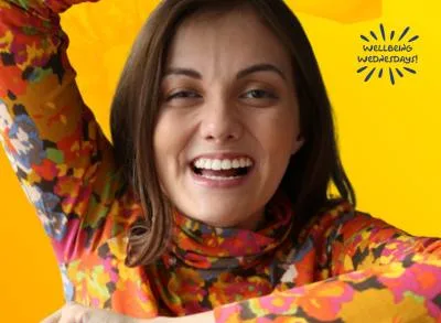 400×293 WW_TAL Tal Anderson is smiling and wearing a colorful flowery turtle neck and holding up her arms against a yellow background.