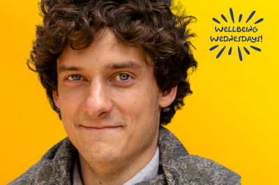 Mar06_BLOG-Wellbeing_Wednesday-Andrew_Arboe (400 x 293 px) Andrew is smiling against a yellow background with a "Wellbeing Wednesday" sticker in navy blue.