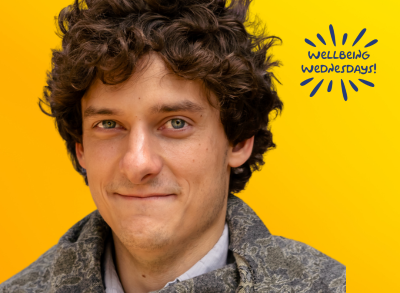 Mar06_BLOG-Wellbeing_Wednesday-Andrew_Arboe (400 x 293 px) Andrew is smiling against a yellow background with a "Wellbeing Wednesday" sticker in navy blue.