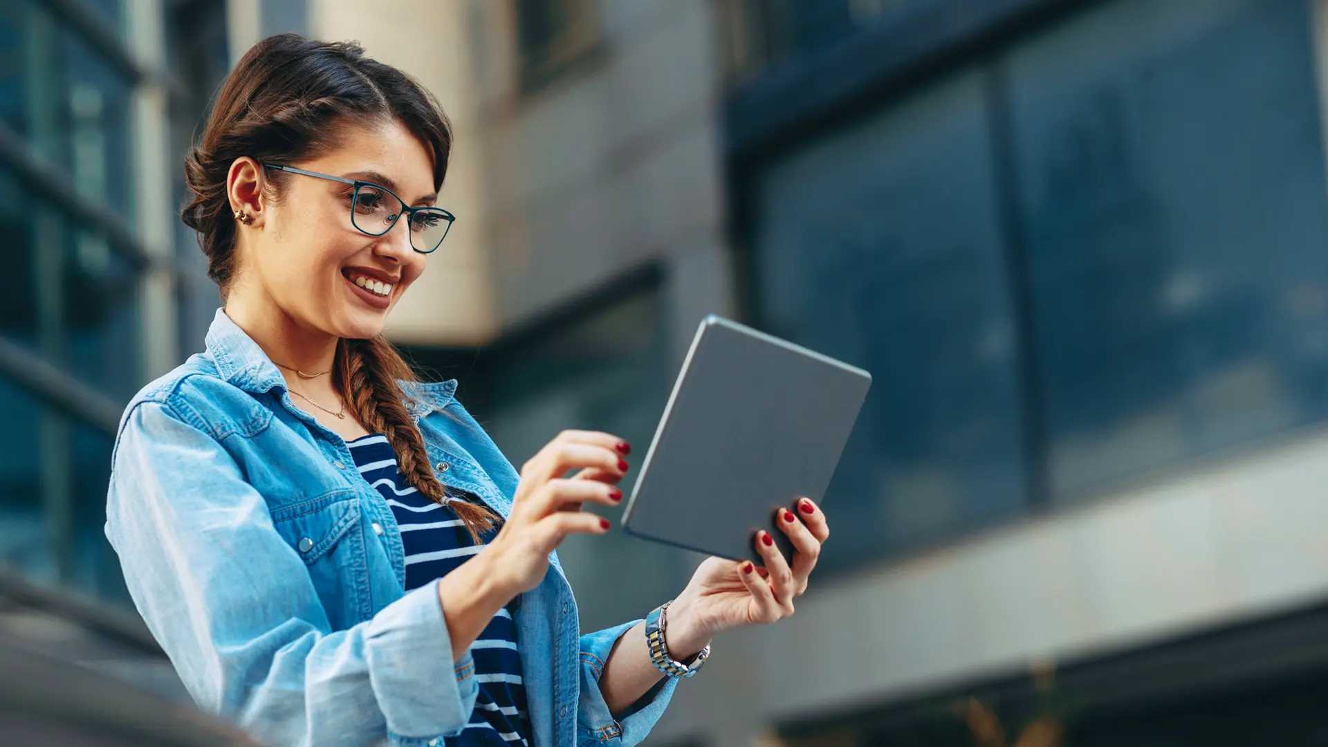 How’s Your Wellbeing? A woman with glasses is outside of a building and looking at her tablet.