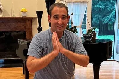 400x293_Yoga A picture of a young man, Ted, doing a prayer yoga pose and smiling. He's wearing a blue shirt and standing in a living room.