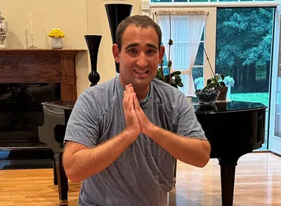 400x293_Yoga A picture of a young man, Ted, doing a prayer yoga pose and smiling. He's wearing a blue shirt and standing in a living room.