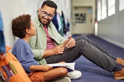400x293_KindnessDay Male teacher sitting on the floor in a hallway with a student who has a journal open. The student and the teacher are smiling.