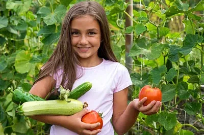 400x293_NatureBlog_GardenGirl A young girl wearing a pink tshirt standing in front of a garden holding tomatoes and zucchini and smiling.
