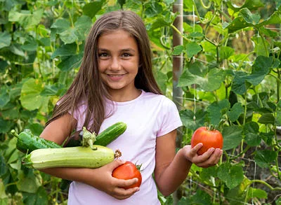 400x293_NatureBlog_GardenGirl A young girl wearing a pink tshirt standing in front of a garden holding tomatoes and zucchini and smiling.