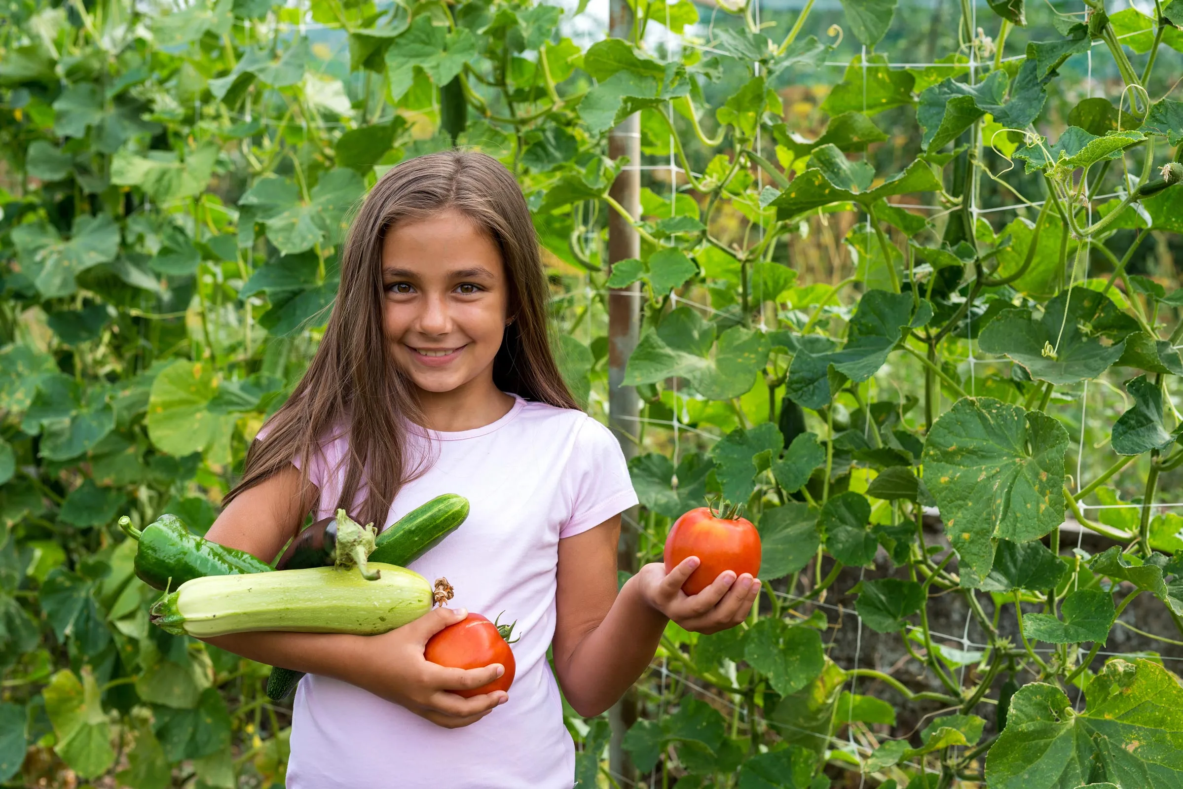 a girl smiling standing in front of a garden holding fruits and vegetables