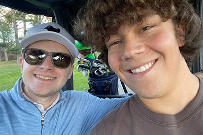 400x293_Noah Noah and Sebastian smile while sitting in a golf cart. Sebastian is in sunglasses, a hat, and blue sweater and Noah is in a brown t-shirt.