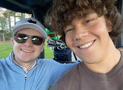 400x293_Noah Noah and Sebastian smile while sitting in a golf cart. Sebastian is in sunglasses, a hat, and blue sweater and Noah is in a brown t-shirt.