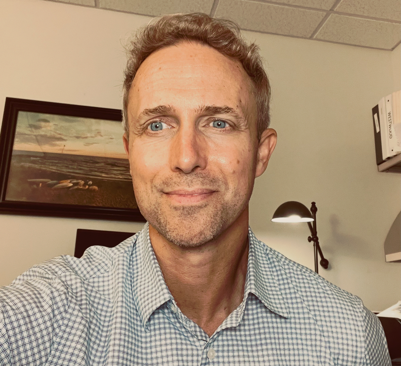 67N267cg Smiling man with blue eyes and short gray hair taking a selfie in an office, wearing a checkered shirt.