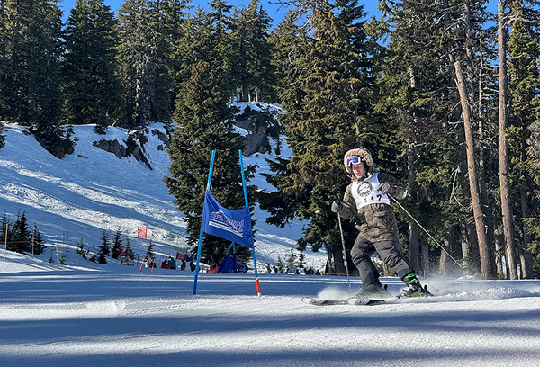 Skier wearing bib number 12 racing down a snowy slope between blue gate markers with forested mountains in the background.
